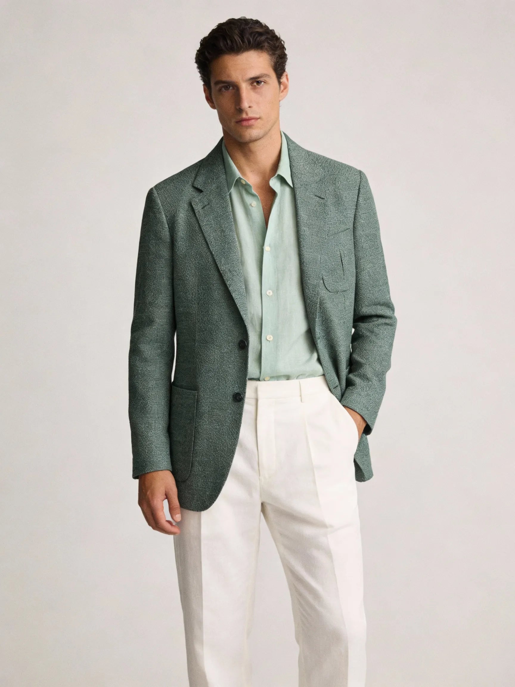 Man wearing a green blazer over a light green shirt and white pants on a plain background